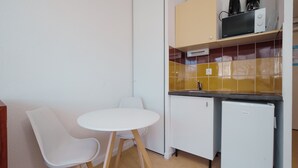 Eine kompakte Küchenzeile mit einem weißen Kühlschrank, einer kleinen Spüle und einer Kaffeemaschine auf der Arbeitsplatte. Ein weißer Tisch mit zwei Stühlen und eine gelb geflieste Spritzschutzleiste sind ebenfalls vorhanden.