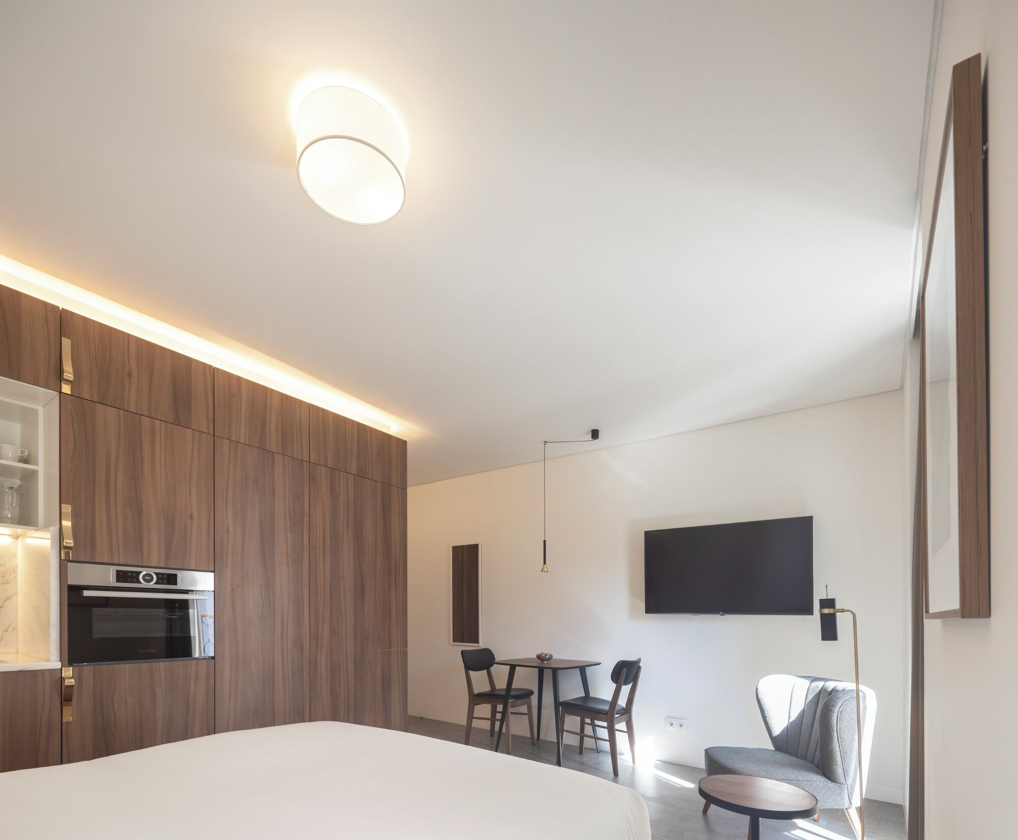A modern hotel room with a large bed, a built-in oven, a dining area with a table and chairs, and a flat-screen TV mounted on the wall.