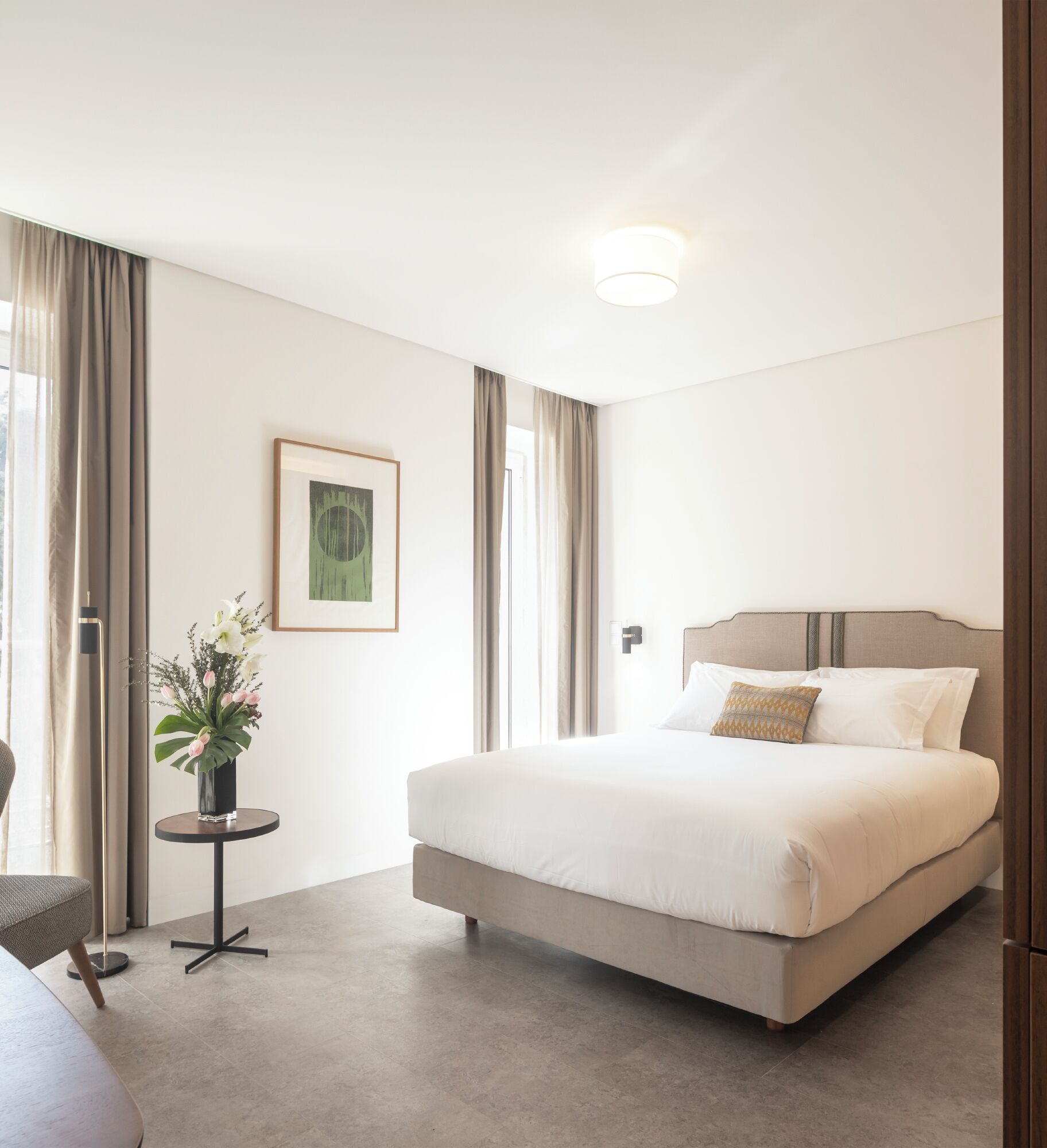 A modern hotel room with a large bed, a small round table with a vase of flowers, and a framed picture on the wall.
