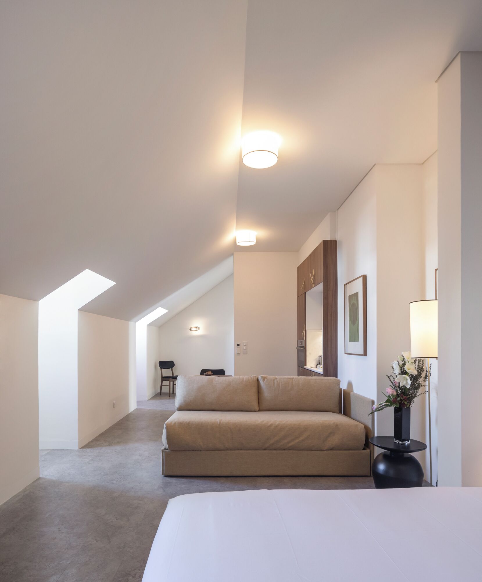 A modern hotel room with a large bed, a sofa, a small table with a vase, and a desk with a lamp. There are skylights on the ceiling, a chair, and a framed picture on the wall.