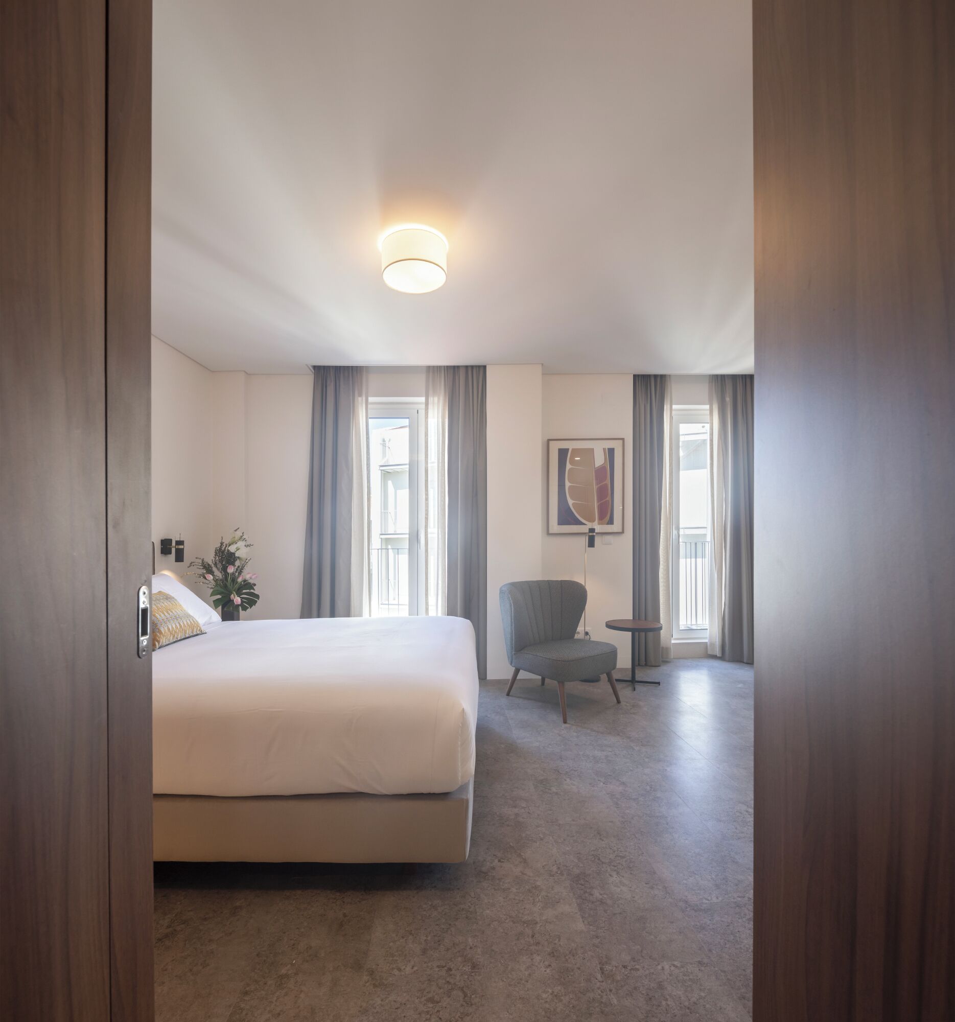 A hotel room with a bed, a chair, a small table, and a painting on the wall.