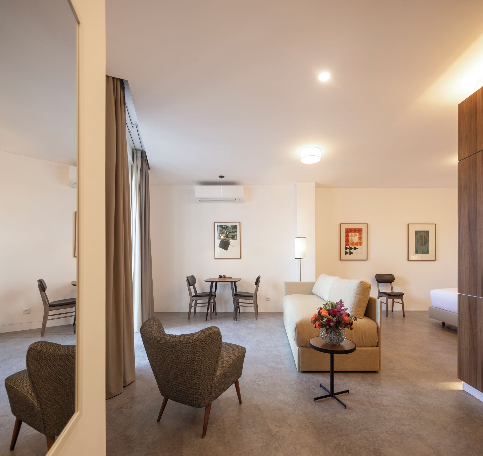 Ein modernes Hotelzimmer mit Sofa, Sessel, Esstisch und Stühlen. Ein kleiner Beistelltisch mit Blumenvase steht daneben.
