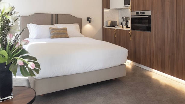 A modern bedroom with a large bed, a bedside table with a vase of flowers, a built-in kitchen with appliances, and a wooden panel wall. - LSA Avenida by Numa (Lisbon)