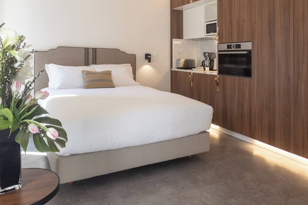 Een moderne slaapkamer met een groot bed, een nachtkastje met een bloevas, een geïntegreerde keuken met apparatuur, en een wand van houten panelen.