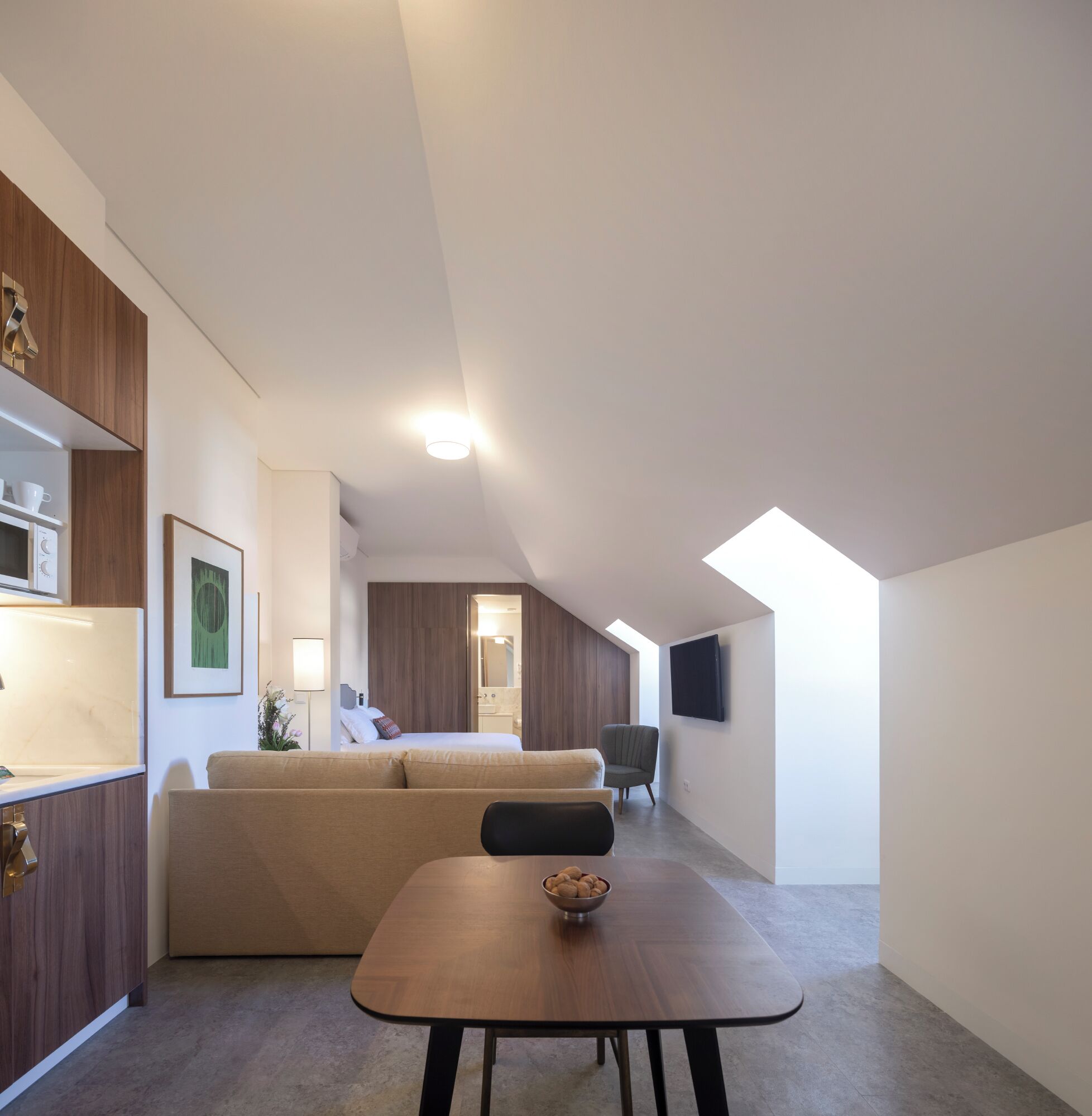 Een moderne, compacte leefruimte met een kitchenette, een bank, een eettafel en een aan de muur bevestigde televisie.