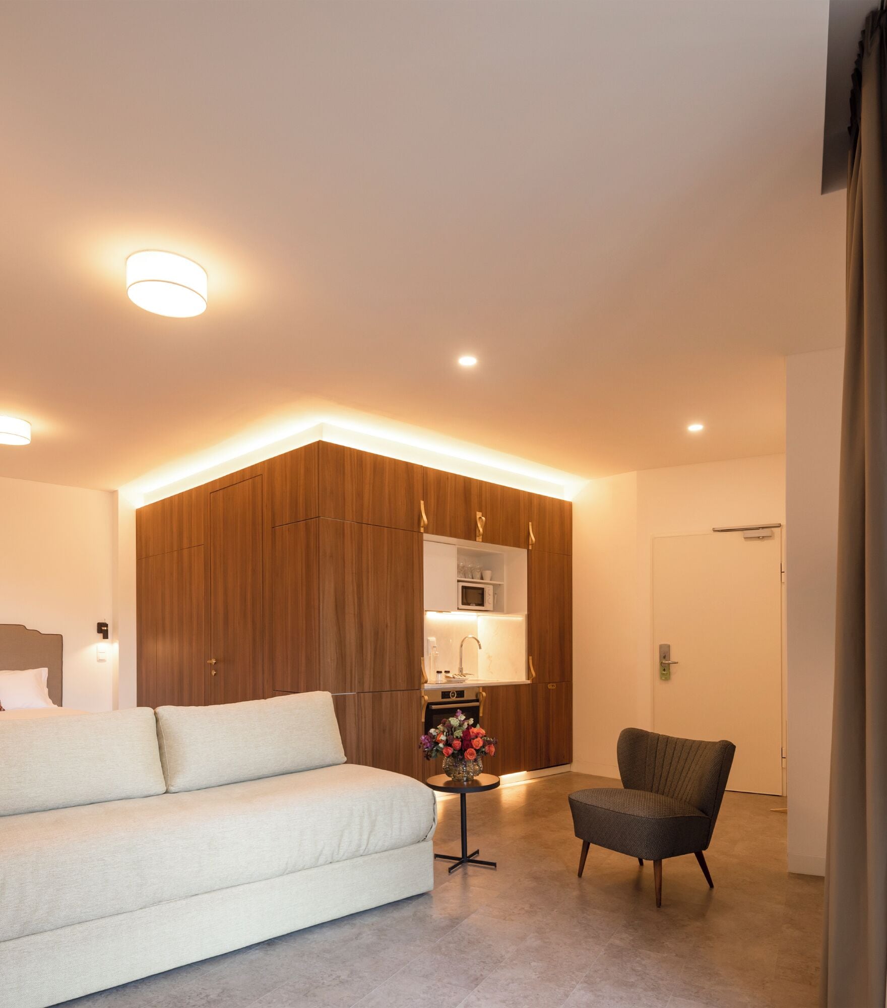 A modern hotel room with a large bed, a seating area, a kitchenette, and a bathroom.