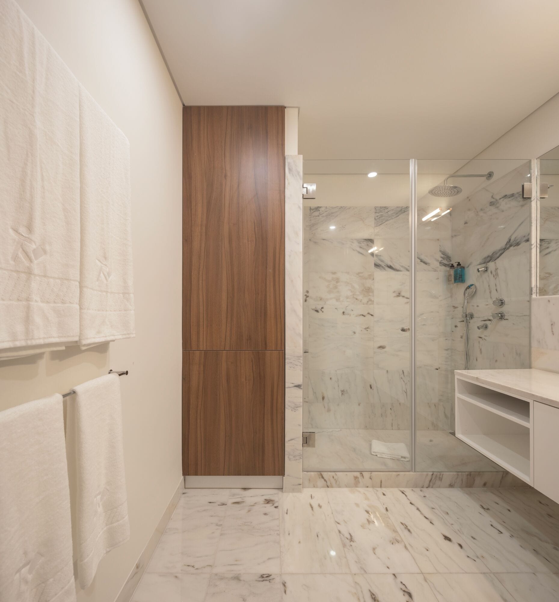 Een moderne badkamer met een houten paneel, marmeren tegels, een glazen douchecabine en een witte wastafel.