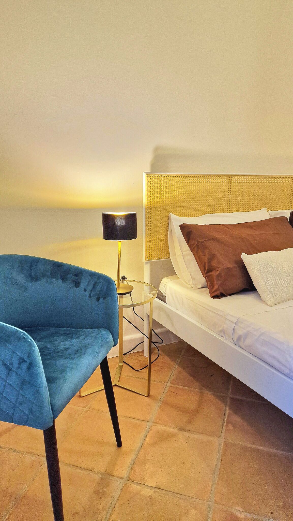 A room with a bed, a blue chair, a small table with a lamp, and a view of the sea.