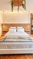 Uma cama com cabeceira de madeira, duas mesas de cabeceira, uma planta pendente e um luminário fixado na parede.