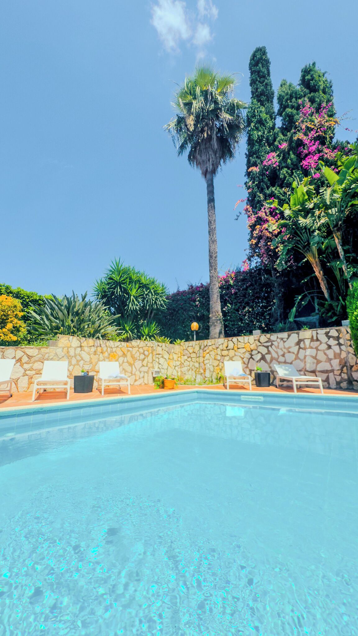 A clear swimming pool with lounge chairs and a stone wall.