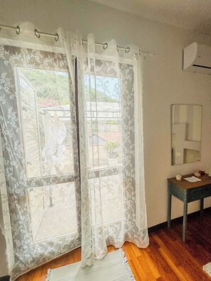Ein Zimmer mit großem Fenster, das mit durchsichtigen Vorhängen drapiert ist, Holzboden, ein kleiner Tisch mit Spiegel und eine Wand-Klimaanlage.