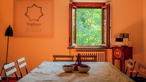 A dining area with a table set for two, a window with a view of greenery, a radiator below the window, and a wall-mounted sign with the text "YogAyur naturale benessere". - Podere tra gli Ulivi (Parrano)