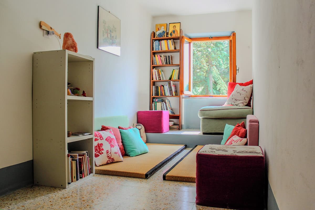 A cozy reading nook with a bookshelf, a cushioned bench, and a window with a view of trees.