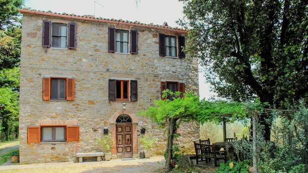 A stone building with a central entrance, multiple windows with shutters, and a garden area with a bench and table. - Podere tra gli Ulivi (Parrano)