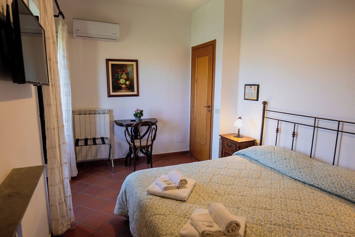 A bedroom with a bed, a bedside table with a lamp, a chair, a small table, and a painting on the wall.