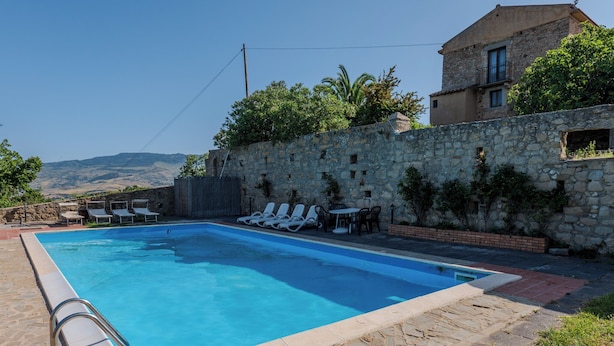 A swimming pool with lounge chairs, a stone wall, and a building with a balcony. - Agriturismo Baglio San Pietro (Nicosia)