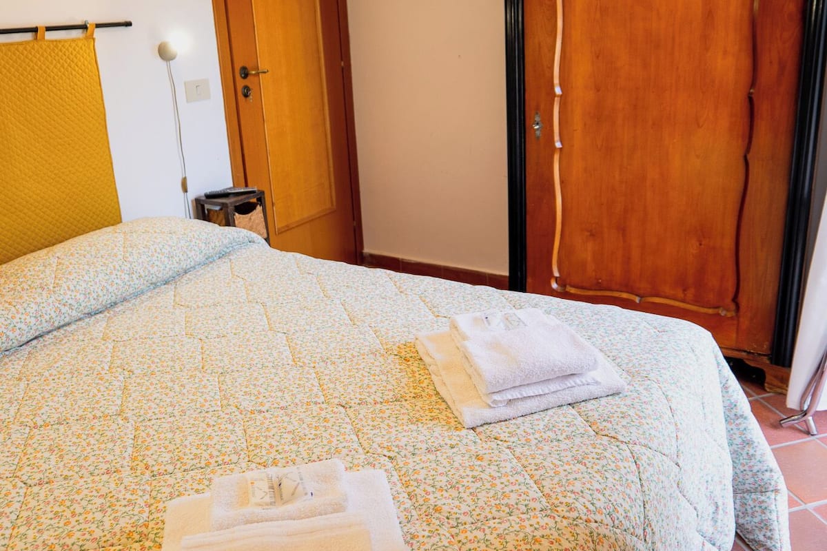 A neatly made bed with a floral bedspread and folded towels, a wooden wardrobe, and a door with a curtain.