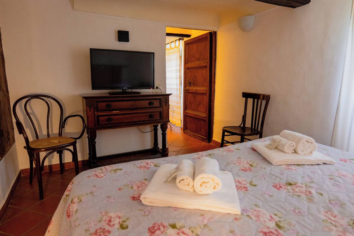 A hotel room with a bed, a television on a dresser, two chairs, and a door leading to another room.