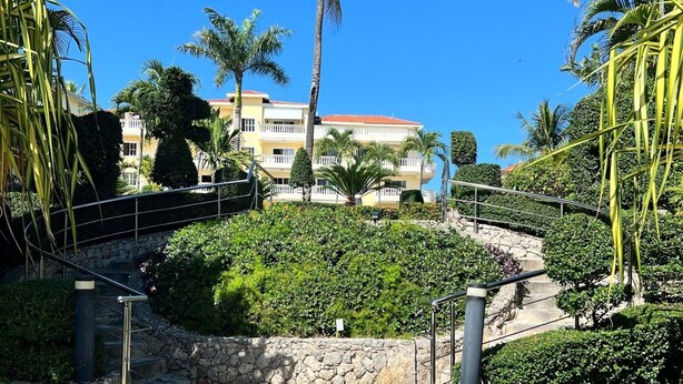 Uma trilha de pedra leva a um edifício com telhado de telhas vermelhas, cercado por vegetação exuberante e palmeiras.