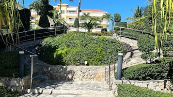 A stone pathway leading to a building with a red-tiled roof, surrounded by lush greenery and palm trees.
