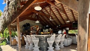Uma área de bar ao ar livre com um telhado de palha, móveis de vime branco e balcão de pedra.