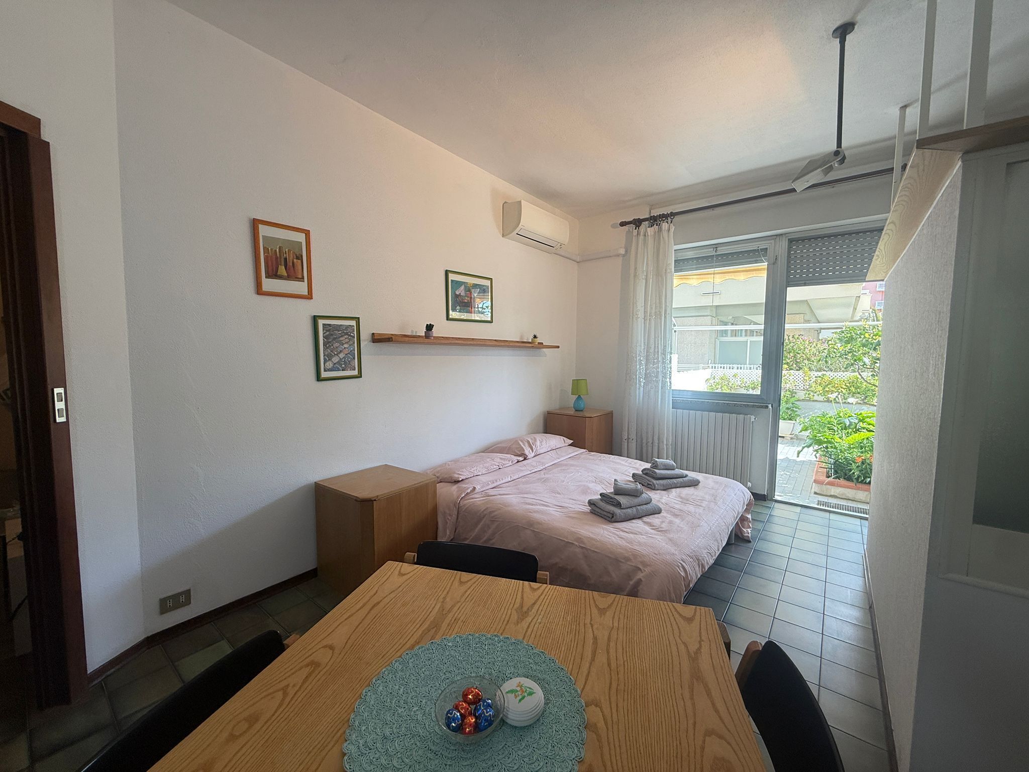 A bedroom with a bed, a bedside table, a window with curtains, a small balcony, and a dining area with a table and chairs.