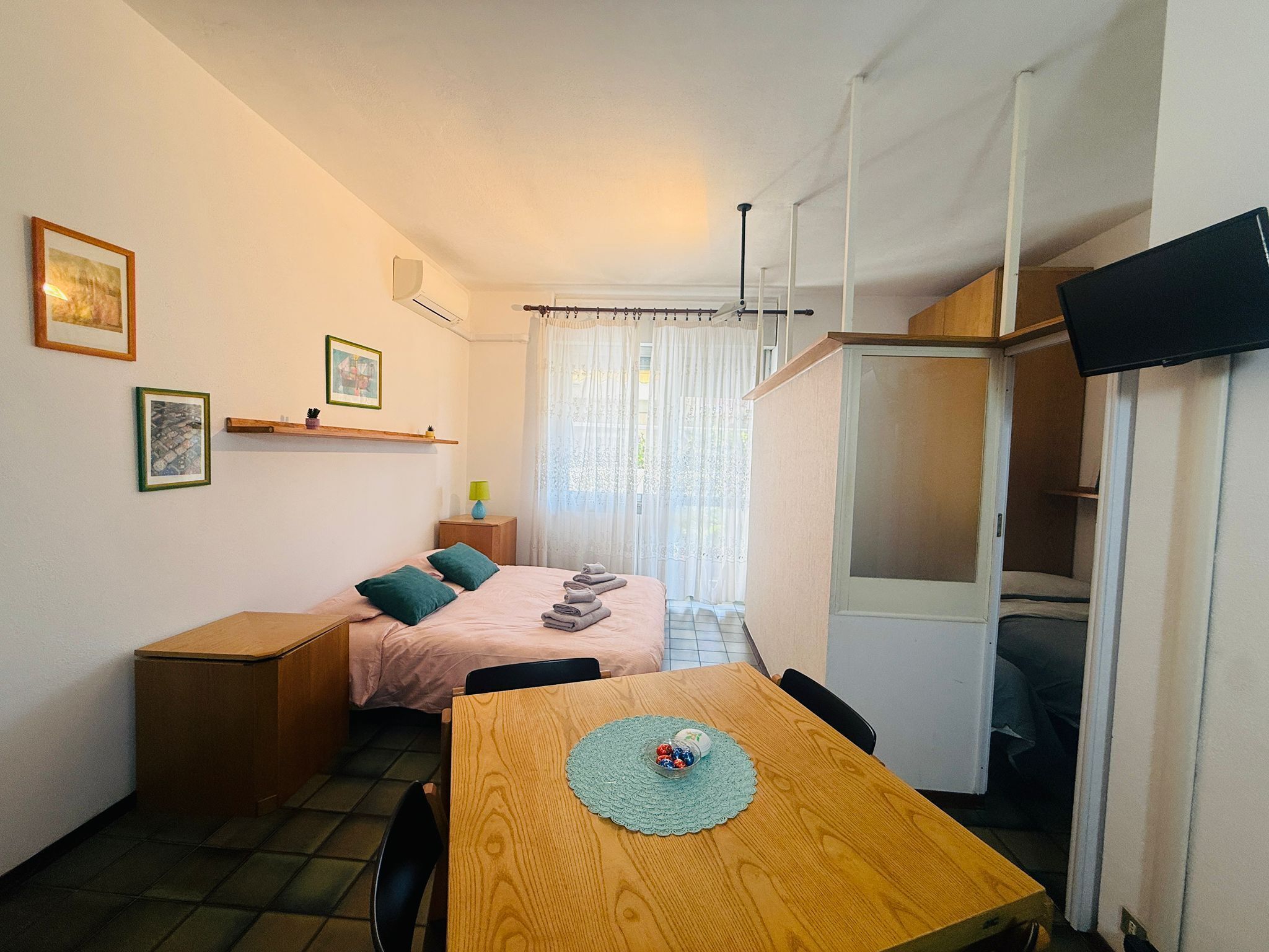 A compact room with a bed, a wooden table with a placemat, a television, and a window with curtains.