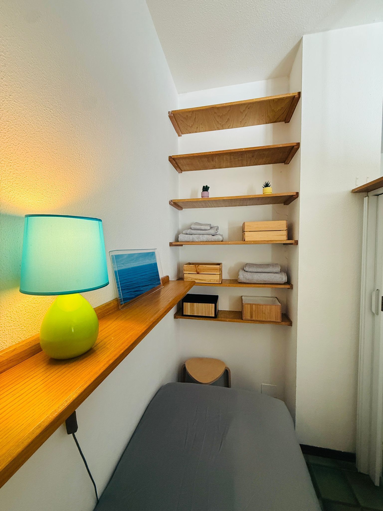 A small, modern room with a wooden desk, a lamp, a bed, and built-in shelves with towels and boxes.