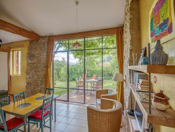 Una habitación con una mesa de comedor, sillas, un sillón de mimbre, una estantería y vistas a un jardín a través de amplios ventanales.