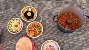 Diversos pratos, incluindo um pote de ensopado, saladas em tigelas e um pão achatado sobre um tapete tecido.