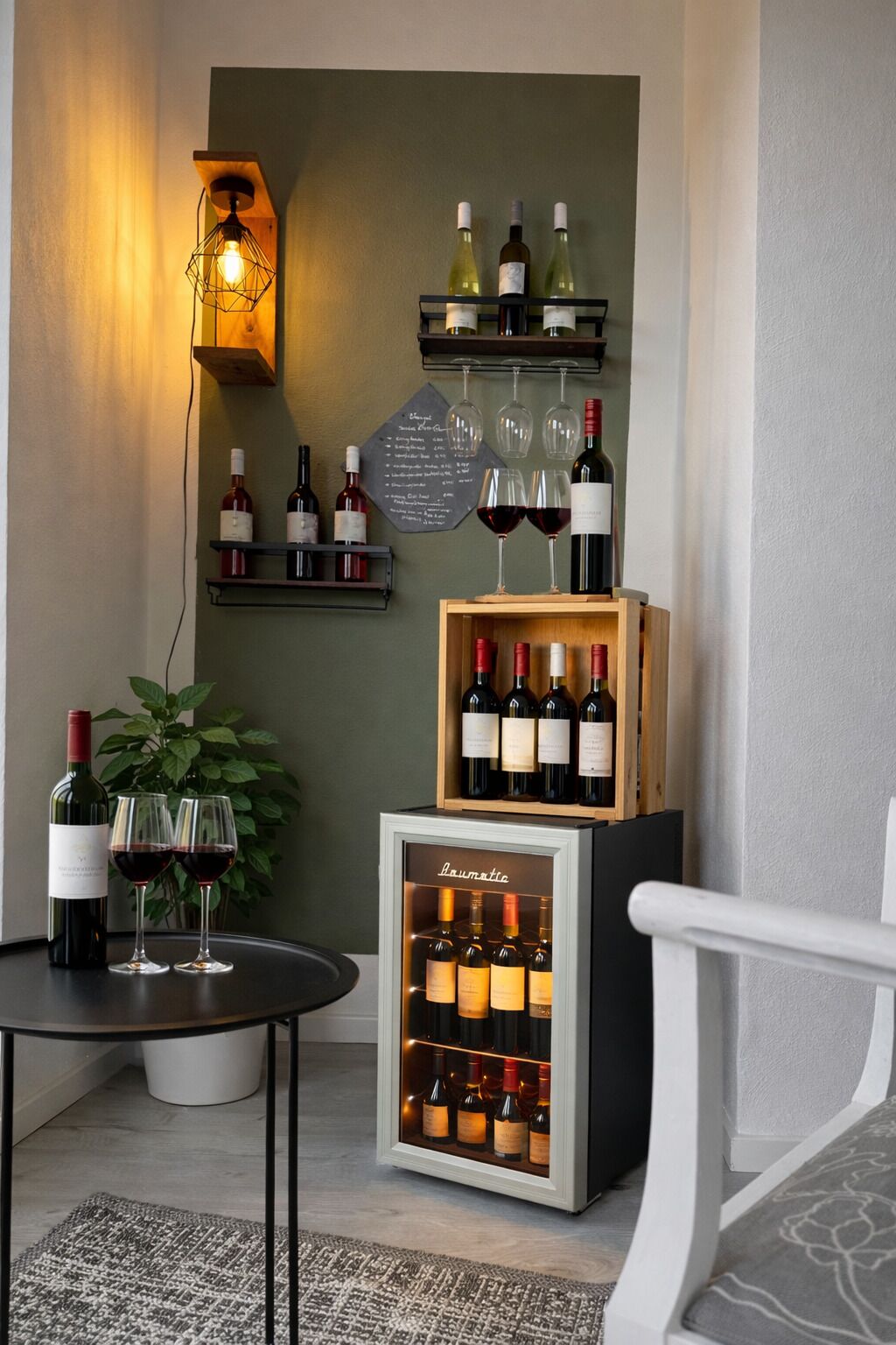 Eine Weinauslage mit Flaschen und Gläsern, ein kleiner Tisch mit einer Flasche und zwei Gläsern sowie ein Kühlschrank mit Weinflaschen.