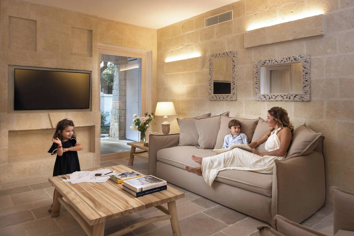 Un soggiorno con un divano color beige, un tavolino in legno con libri, una TV a parete e un muro in pietra.