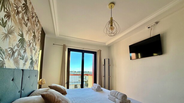 A hotel room with a large bed, a TV mounted on the wall, and a view of the city through the window. - Le Mistral - Elegant apartment with sea view! (Le Grau-du-Roi)