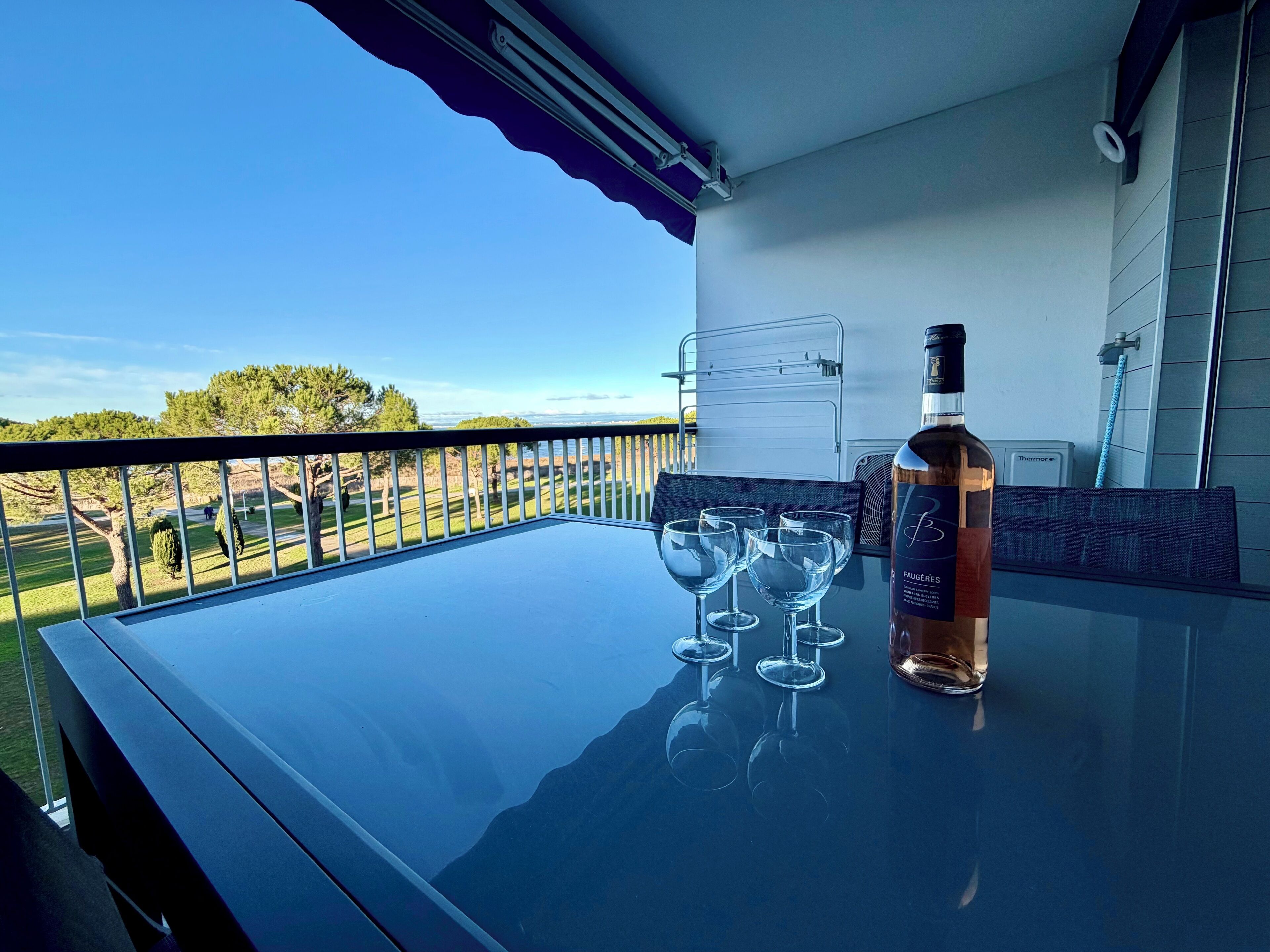 Un balcon avec une table en verre, une bouteille de vin de Faugères et quatre verres à vin.