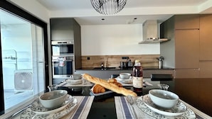 A modern kitchen with a dining area, featuring a large window, stainless steel appliances, and a dining set with bread and a bottle of wine. - Le Mistral - Elegant apartment with sea view! (Le Grau-du-Roi)