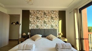 A hotel room with a large bed, two nightstands, a wall with a tropical leaf pattern, and a view of the outdoors through a sliding door. - Le Mistral - Elegant apartment with sea view! (Le Grau-du-Roi)
