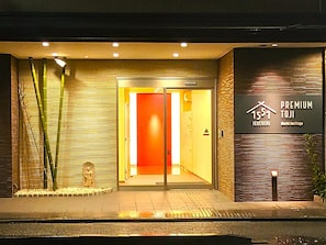 A building entrance with a glass door, a sign reading 'PREMIUM TOJI,' and a bamboo plant to the left.