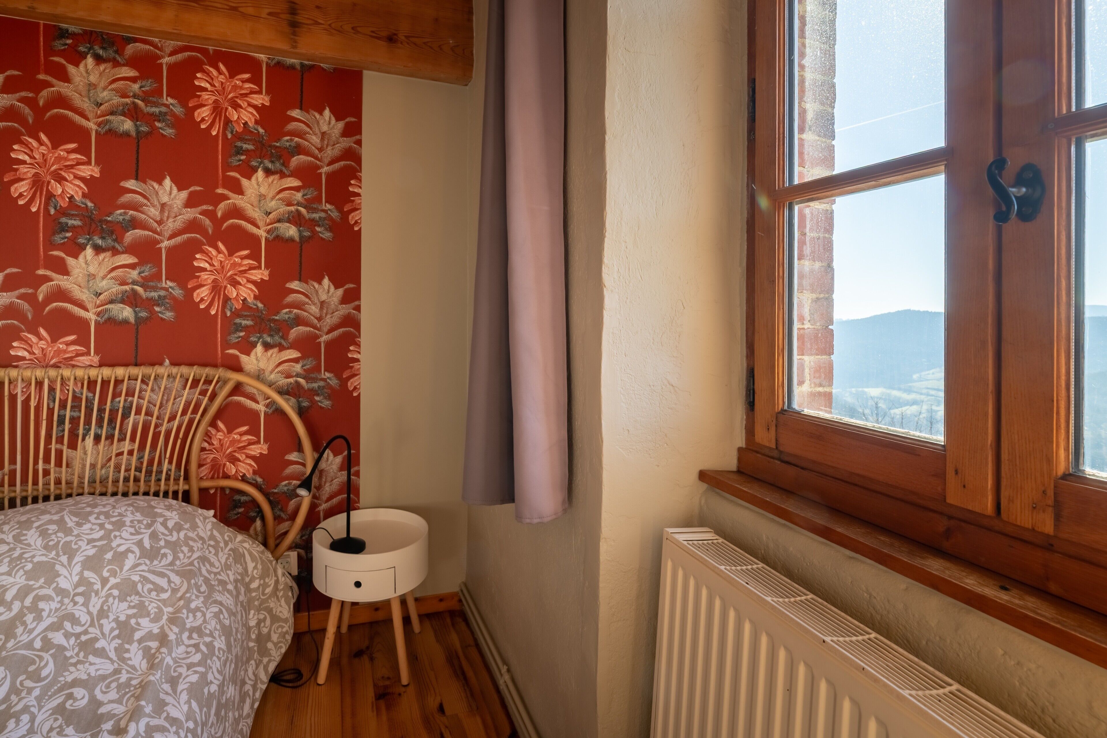 Un dormitorio con una cama, una mesita de noche pequeña, un radiador, una ventana con cortinas y una pared con estampado floral.