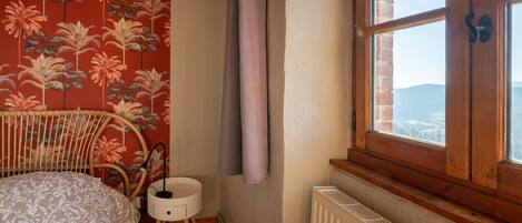 Un dormitorio con una cama, una mesita de noche pequeña, un radiador, una ventana con cortinas y una pared con estampado floral.