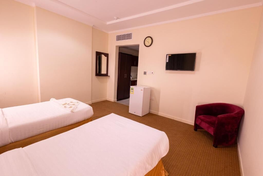Habitación de hotel con dos camas individuales, un escritorio pequeño con una silla, un televisor y un sillón rojo.