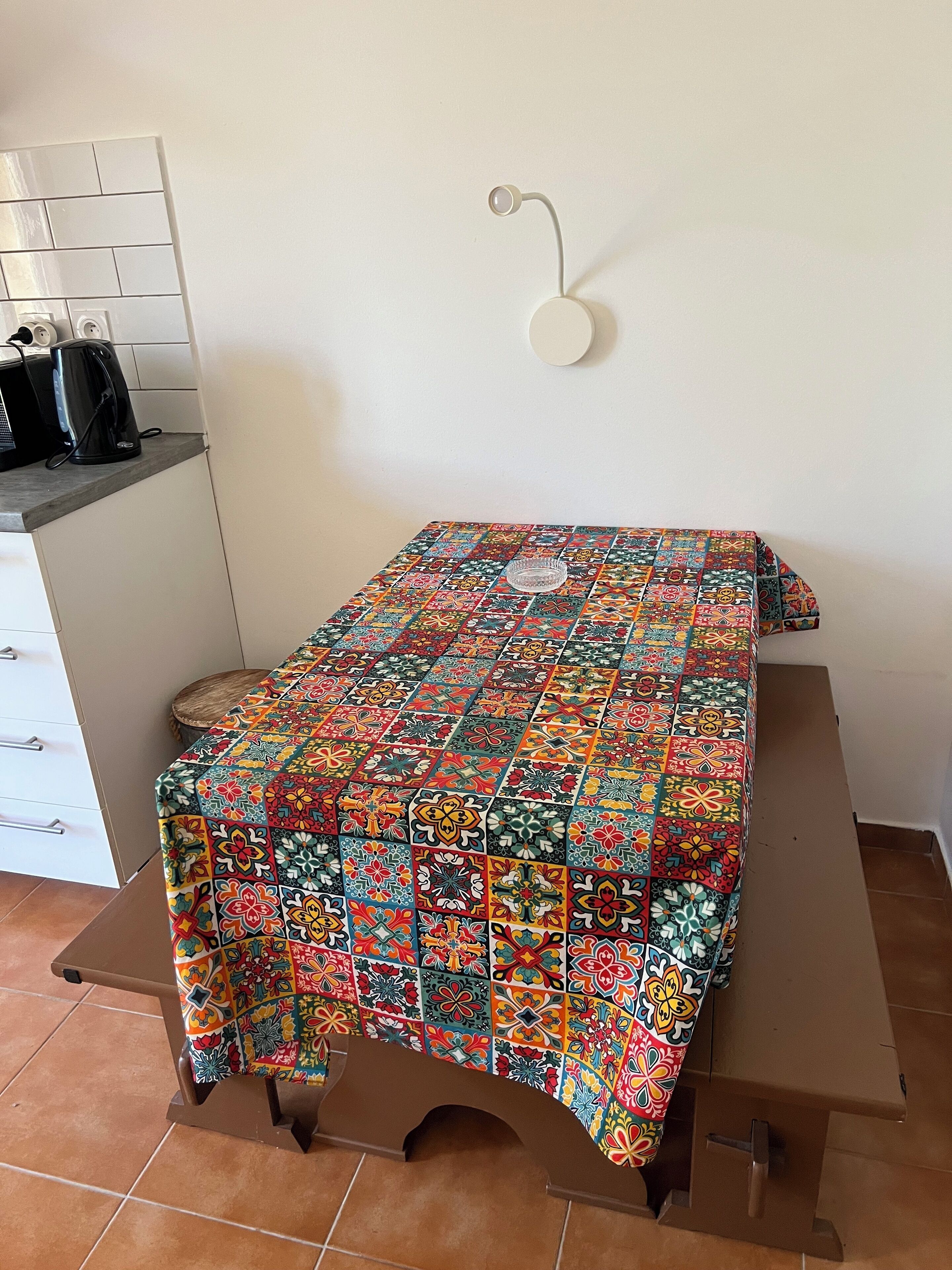 A small, neatly arranged room with a colorful tablecloth-covered table, a wall-mounted light, and a tiled floor.