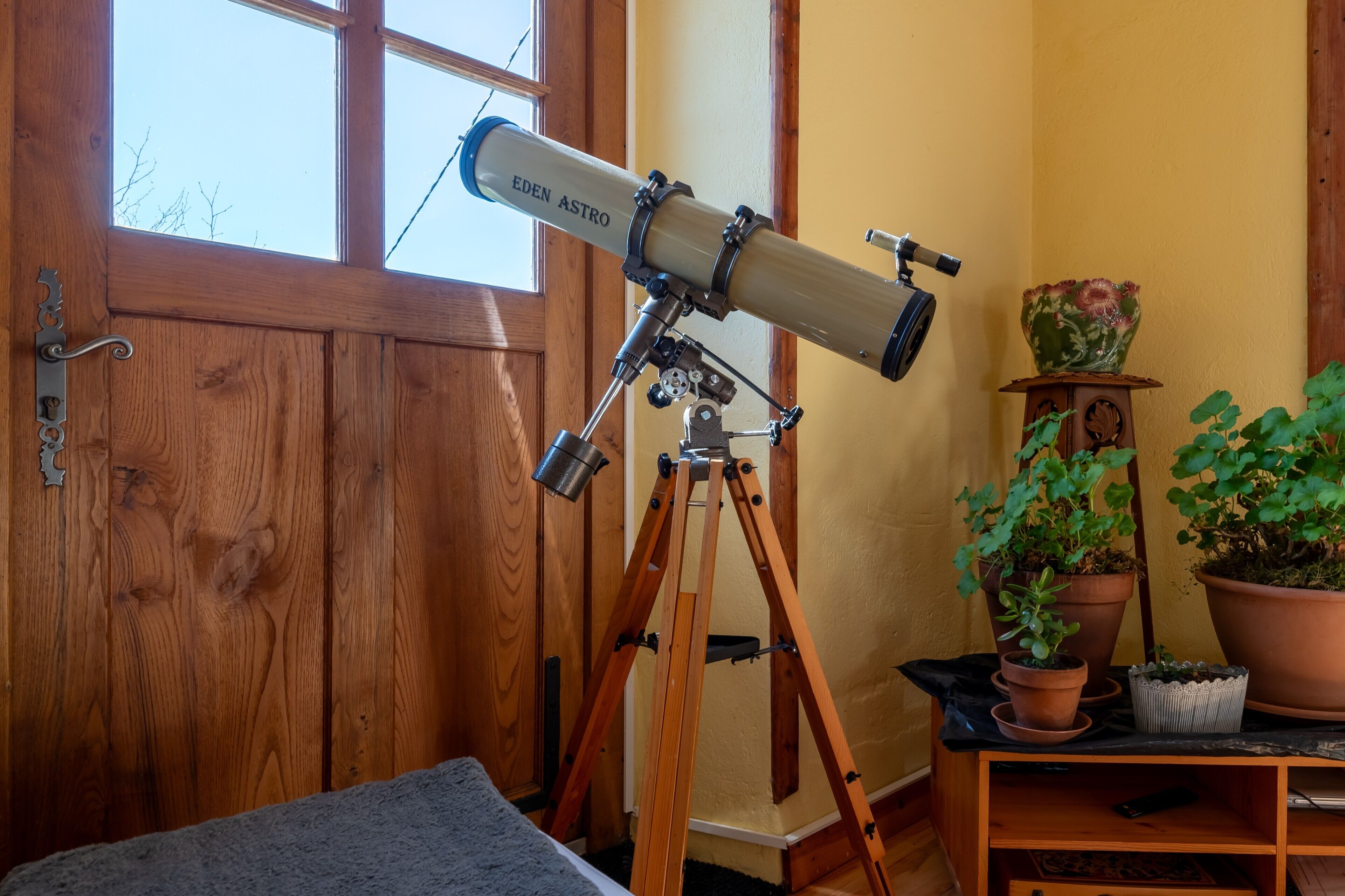 A telescope labeled 'EDEN ASTRO' on a wooden tripod stand, positioned near a wooden door and a window.