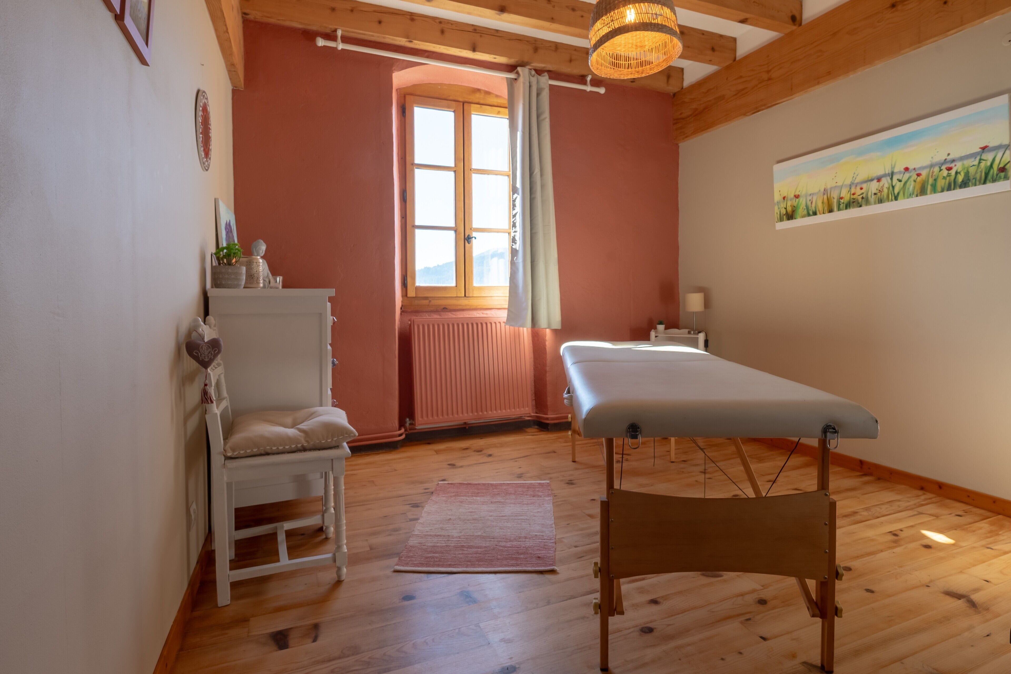 A room with a wooden floor, a white chair, a massage table, a window with curtains, and a painting on the wall.