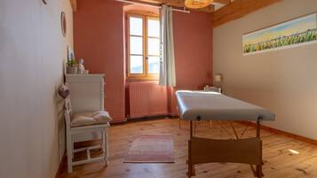 A room with a wooden floor, a white chair, a massage table, a window with curtains, and a painting on the wall.