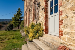 Un bâtiment en pierre avec une porte blanche, des marches menant à l’entrée et un petit jardin agrémenté d’arbustes.