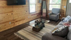 A cozy living room with wooden walls, a flat-screen TV mounted on the wall, a glass coffee table, a grey armchair, and a sofa with patterned cushions. - Sowell’s Cozy Cottage (Milton)