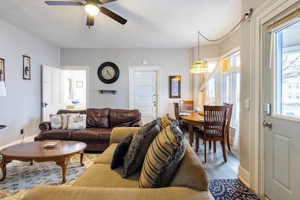 A living room with a brown leather sofa, a beige sofa, a wooden coffee table, a dining table with chairs, a ceiling fan, and a clock on the wall.
