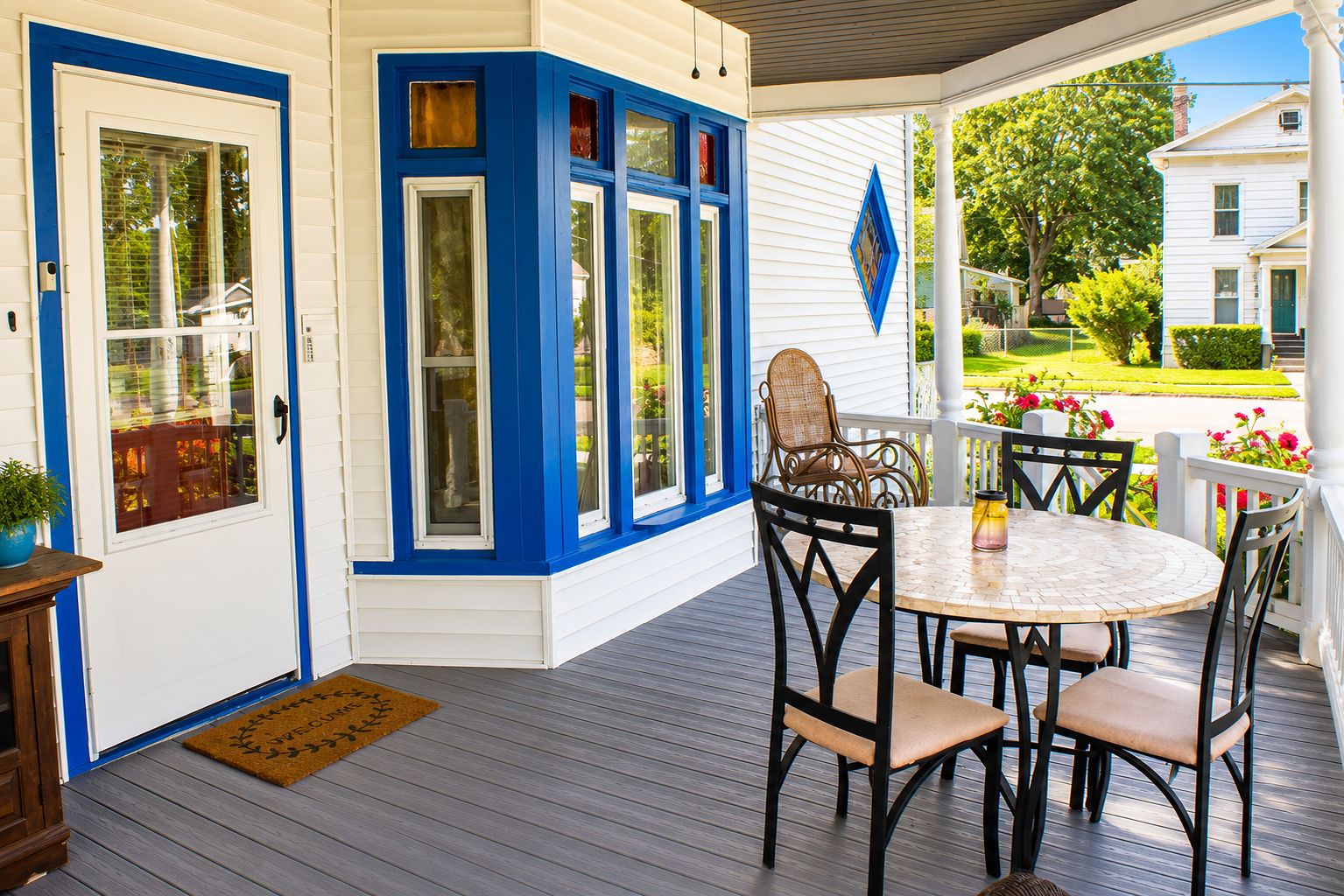 A covered porch with a round table and chairs, a door with blue trim, and a view of a green lawn and white house.