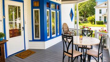 A covered porch with a round table and chairs, a door with blue trim, and a view of a green lawn and white house.