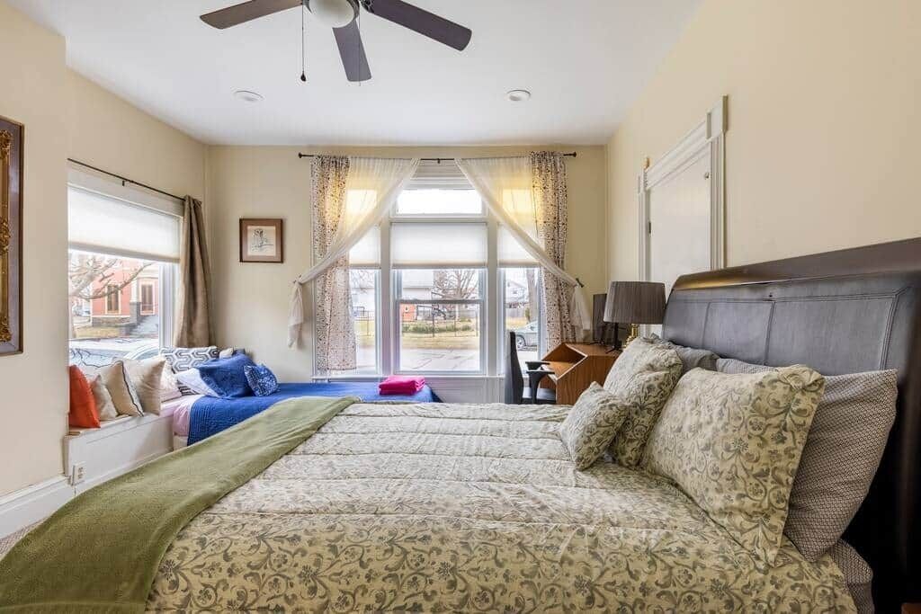 Un dormitorio con una cama grande, un escritorio, una silla, un ventilador de techo y una ventana con cortinas.